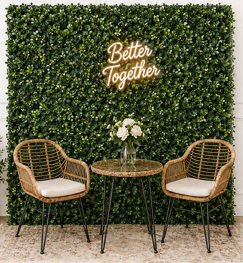 A lush green hedge wall backdrop with a lit sign and rattan bistro chairs